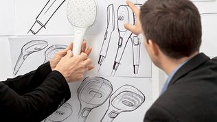 Zeichnungen einer Handbrause an einer Wand vor dem ein Mann steht, und ein anderer eine Handbrause vor einer Zeichnung haelt