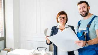 Ein Mann in Arbeitskleidung steht neben einer Frau, die eine Zeichnung in der Hand haelt