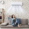 Eine Junge Frau sitzt in einem Raum auf der Couch mit der Fernbedienung für die Klimaanlage die an der Wand über Ihr ist. Aus der Klimaanlage kommt kühle Luft, und die Frau atmet Tief und lächelt.